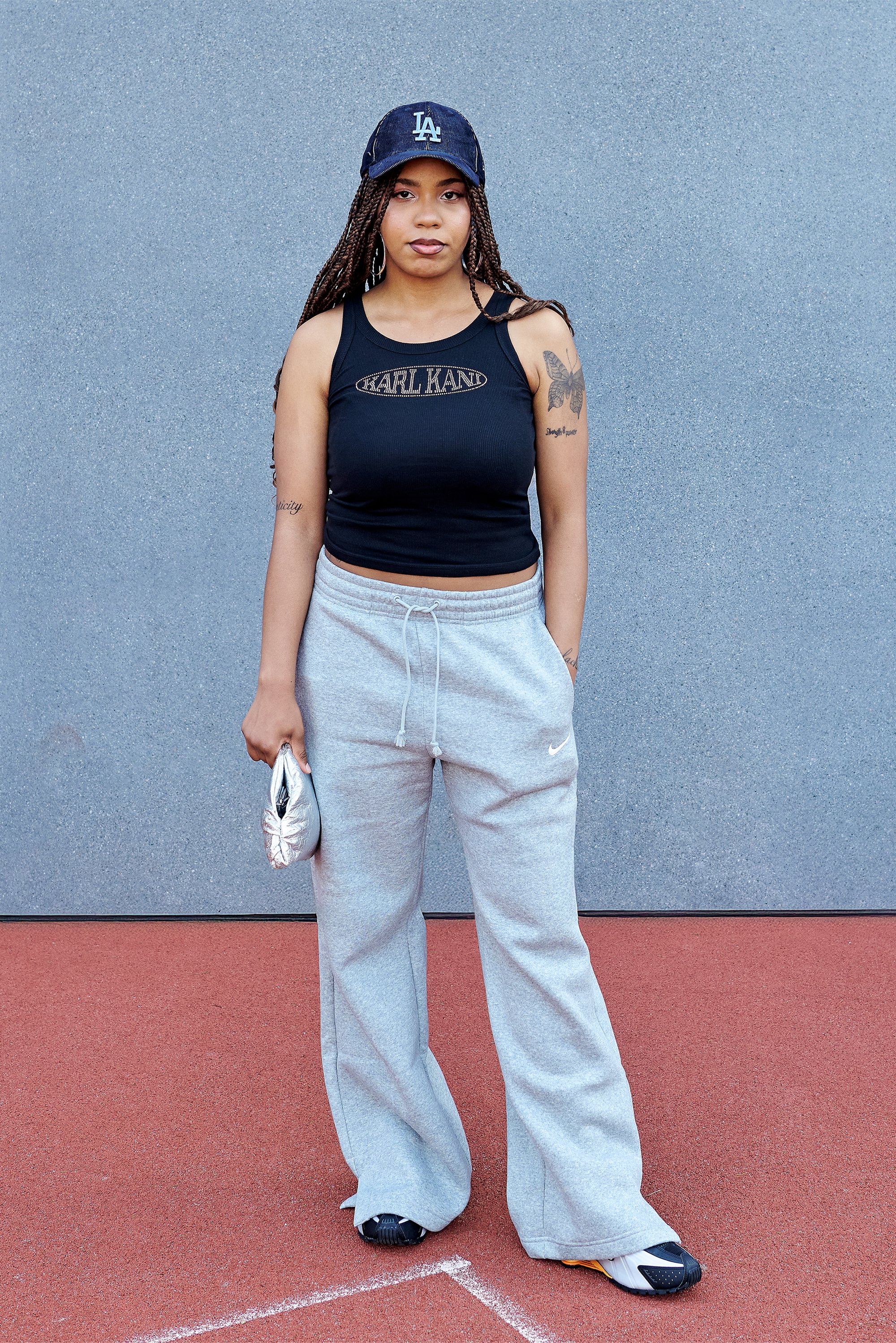Woman with braids wearing a blue LA cap, black tank top, gray sweatpants, and sneakers stands on red track against gray wall, holding silver pouch.