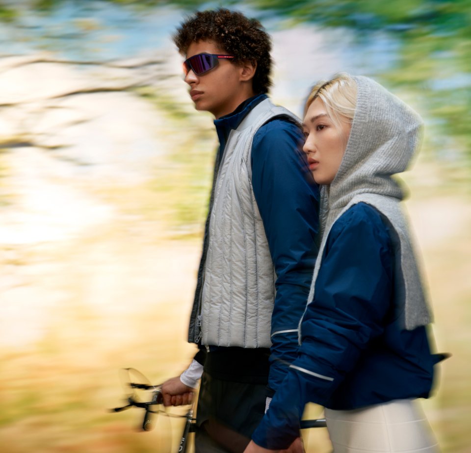 Two young adults stand side by side, dressed in athletic wear, holding bicycle handles, set against a blurred natural background.