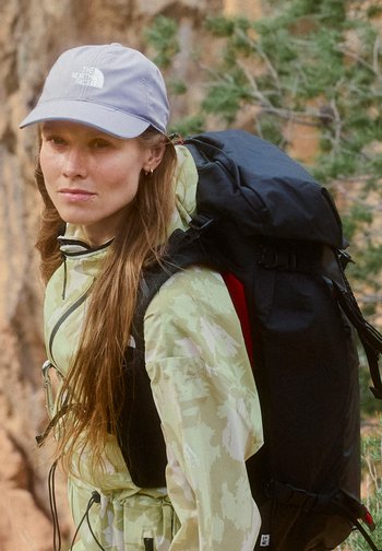Young woman wearing a grey cap and light camo jacket carries a large black hiking backpack outdoors near trees.