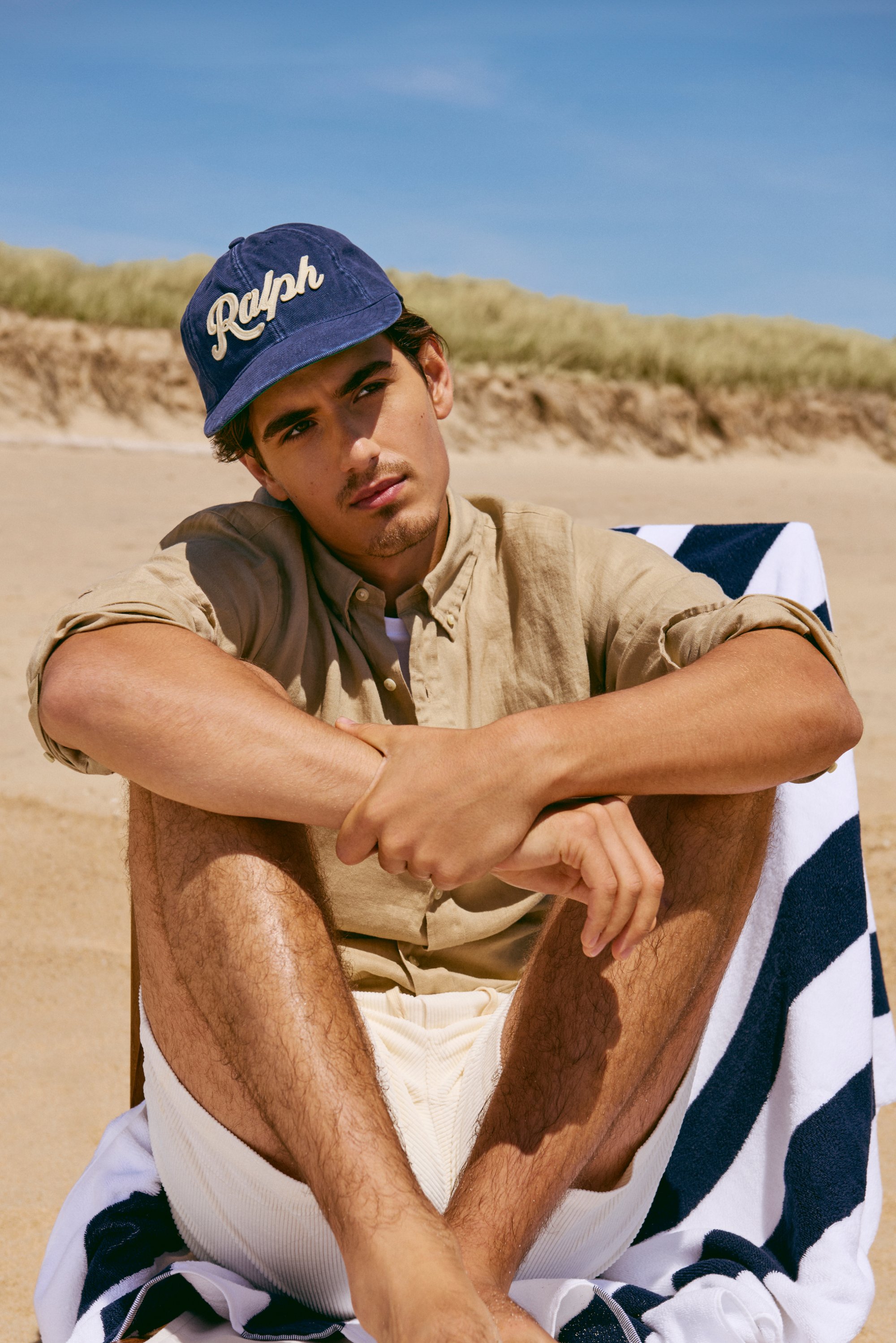 Un joven está sentado en una silla de playa, lleva una gorra azul marino y una camisa clara, con arena y hierba de fondo bajo un cielo azul claro.