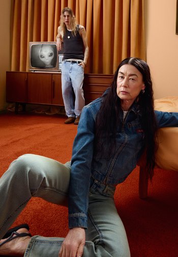 Femme en tenue en denim assise sur un tapis orange près du lit, homme en débardeur noir et jeans s'appuyant sur une armoire avec une télévision vintage affichant un visage d'extraterrestre.