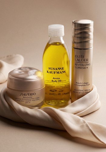Trois produits de soin de la peau sur un tissu beige : crème de jour Shiseido, huile corporelle à l'arnica Susanne Kaufmann et sérum sculptant Estée Lauder.