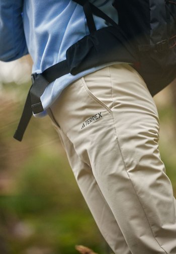 Person som har på seg beige Terrex-bukser og en blå topp med en synlig svart ryggsekkrem, står ute i et naturlig miljø.