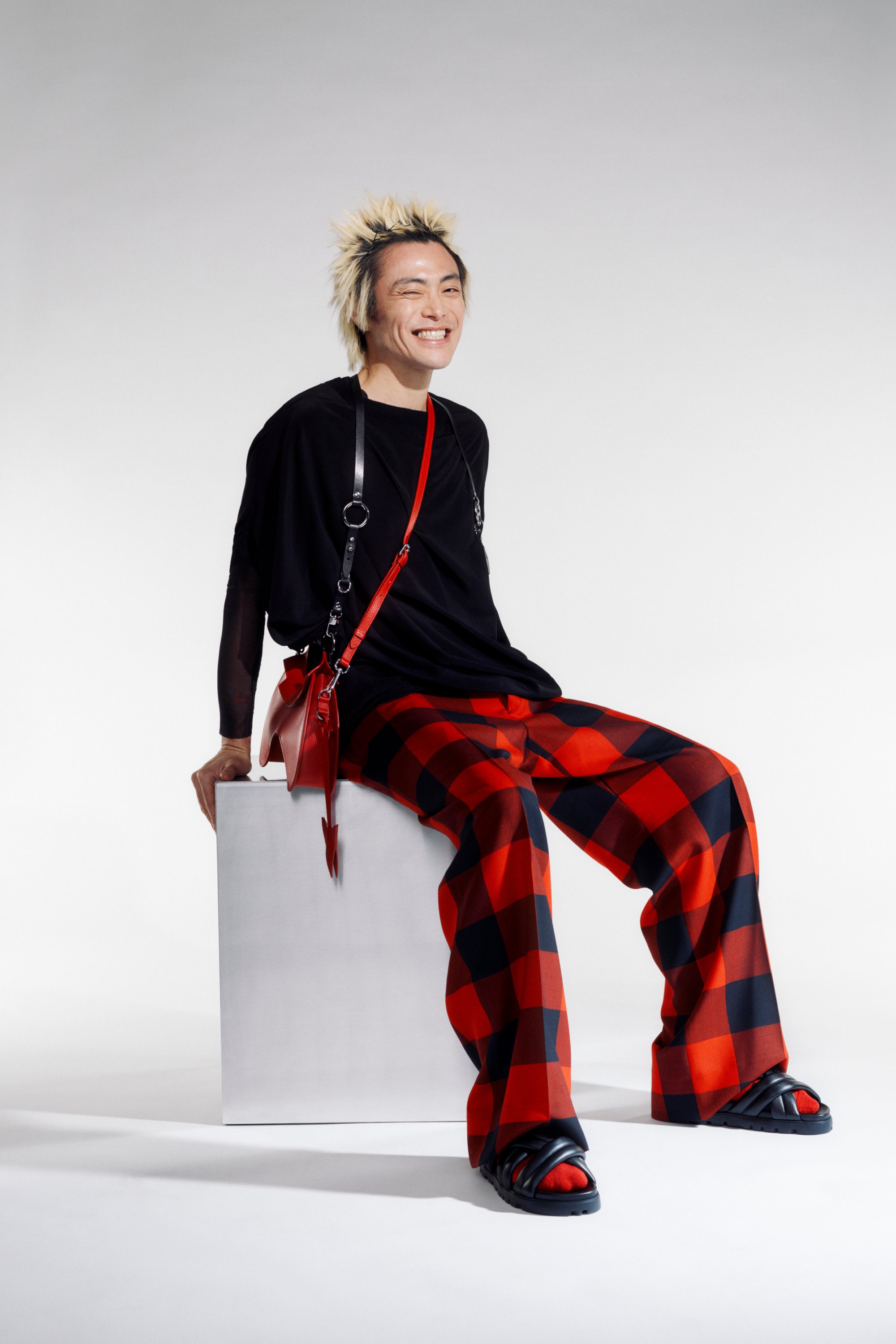 Persona con cabello rubio y puntiagudo sonríe mientras viste una camiseta negra y pantalones a cuadros rojos y negros, sentado sobre un cubo blanco.