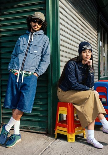 Hombre con chaqueta azul, pantalones cortos y gorra apoyado en una persiana verde; mujer con chaqueta vaquera, falda beige y gorro sentada sobre taburetes apilados en el exterior.