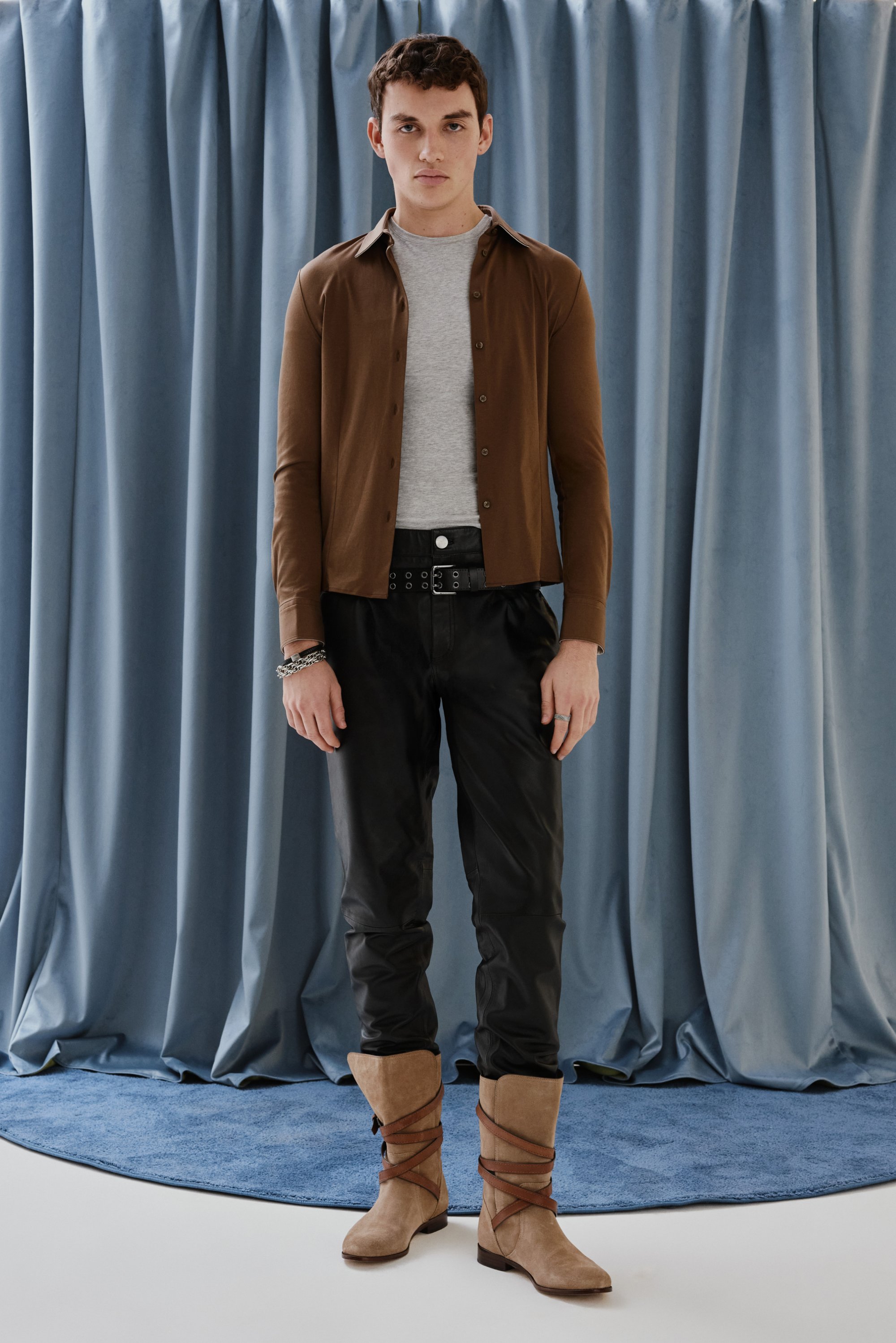 Un joven está de pie sobre una alfombra azul redonda, vistiendo una camisa gris, una chaqueta marrón de botones, pantalones negros y botas beige, frente a cortinas azules drapeadas.