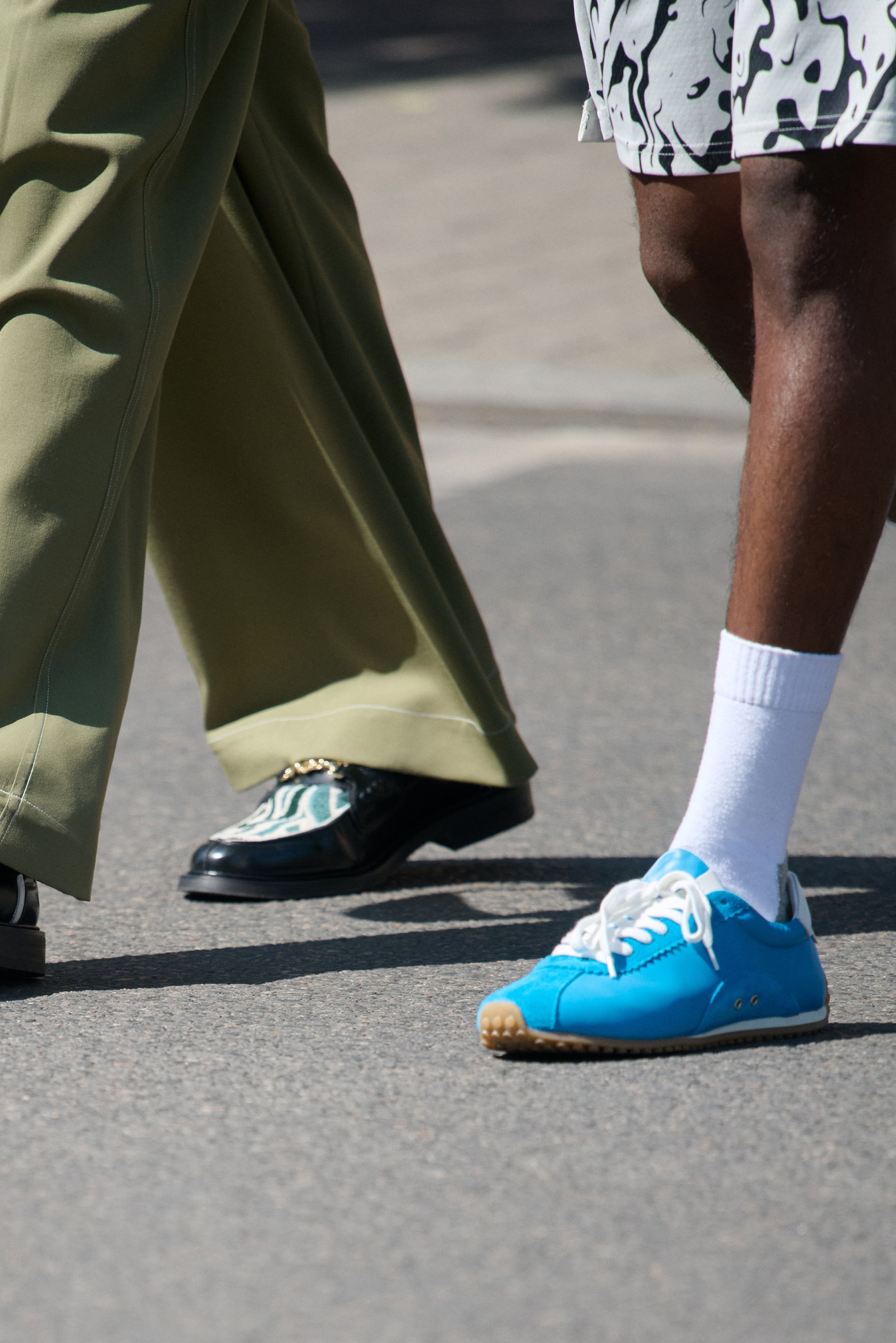 Dos pares de pies caminando sobre el pavimento. Uno lleva un zapato negro con un diseño estampado, el otro una zapatilla azul brillante con cordones blancos.