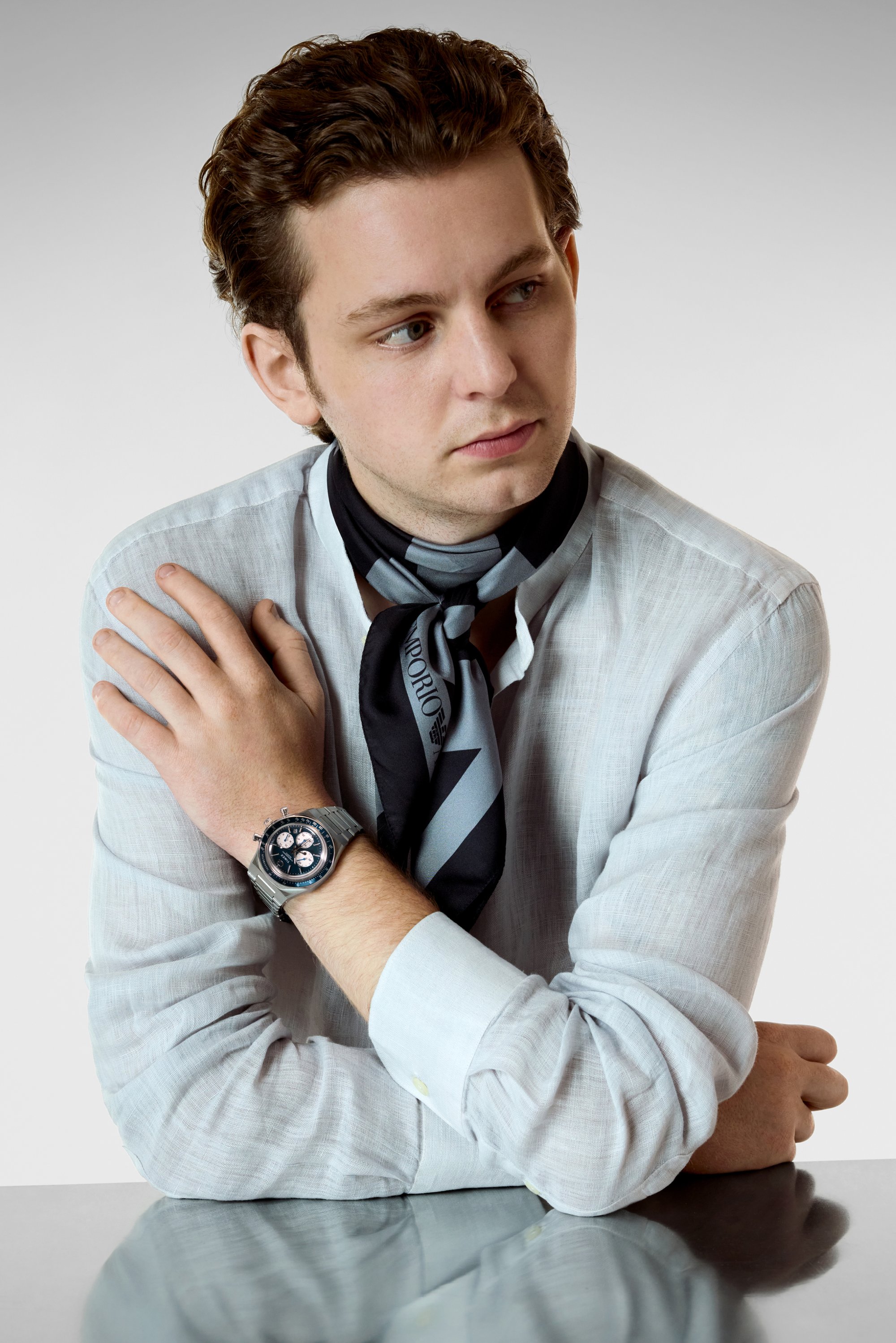 Reloj de pulsera plateado con esfera azul y tres subesferas, usado sobre una camisa azul clara con las mangas arremangadas y una bufanda negra con acentos grises.