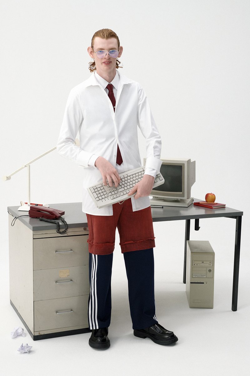 Un joven con una camisa blanca y pantalones cortos rojos sostiene un teclado, de pie al lado de un escritorio con computadoras vintage, una manzana y papeles arrugados.