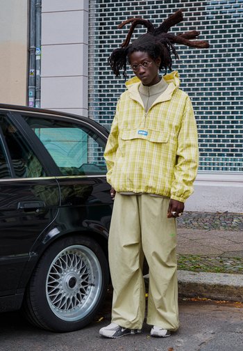 Nagy raszta hajú személy kerek szemüvegben, sárga kockás dzsekiben, bézs nadrágban és sportcipőben áll egy fekete autó mellett egy városi utca közepén.