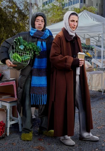 Gevoerde zwarte jas, blauw en bruin gestreepte sjaal, bruine overjas, grijze broek, zachte grijze schoenen; metalen mand met groenten. Marktsetting.