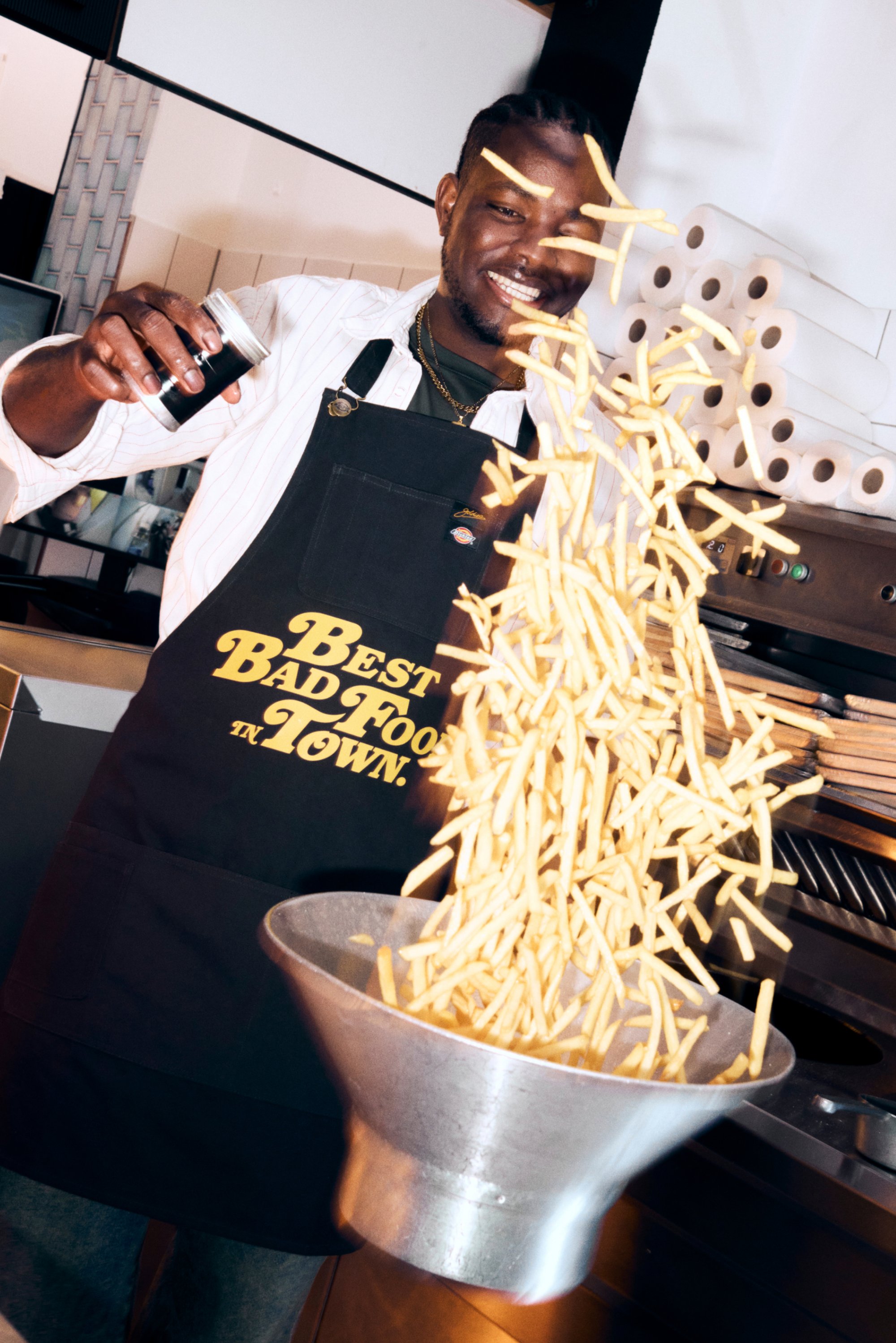 Lächelnder Koch in einer schwarzen Schürze schüttet frisch frittierte Pommes Frites in einen Metallbehälter, während im Hintergrund Küchenausrüstung und Papierrollen zu sehen sind.