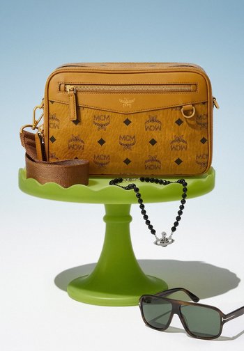 Tan MCM crossbody bag with brown strap displayed on a green stand, black beaded necklace with silver pendant draped over the stand, and black sunglasses placed nearby.
