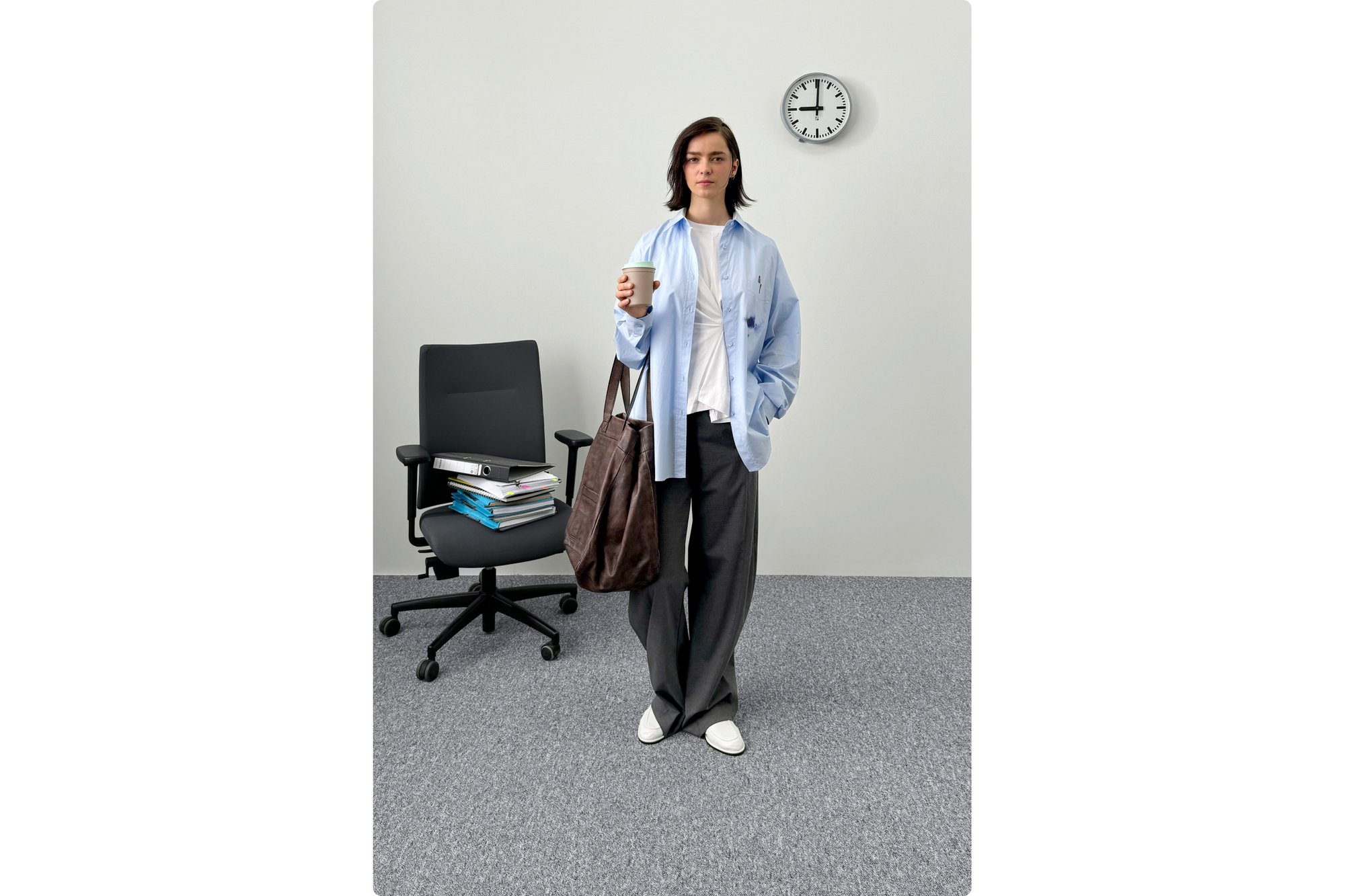 A person with shoulder-length dark hair holds a coffee cup and a brown bag, standing near an office chair and a clock on a plain wall.