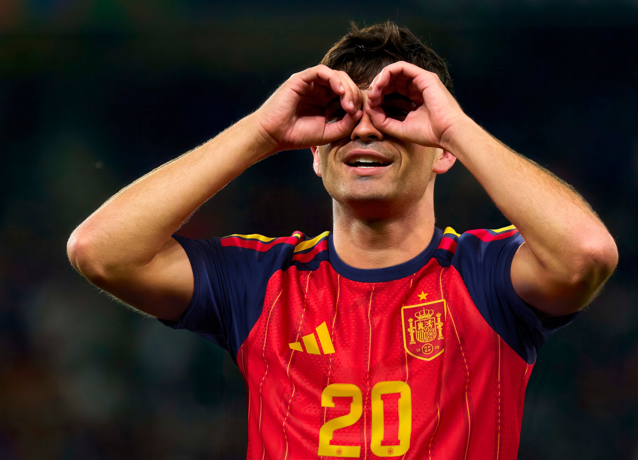 Κόκκινη και μπλε φανέλα ποδοσφαίρου με κίτρινες λεπτομέρειες, με τον αριθμό 20 και έμβλημα. Άτομο που κάνει χειρονομία με τα χέρια κοντά στο πρόσωπο.