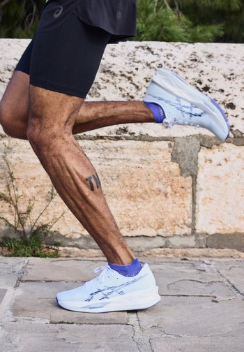 Άτομο που τρέχει έξω φορώντας λευκά αθλητικά παπούτσια και μπλε κάλτσες, με ένα ορατό τατουάζ στη αριστερή γάμπα, με φόντο έναν πέτρινο τοίχο.