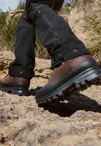 Person wearing black trousers and brown hiking boots walking on rocky outdoor terrain.