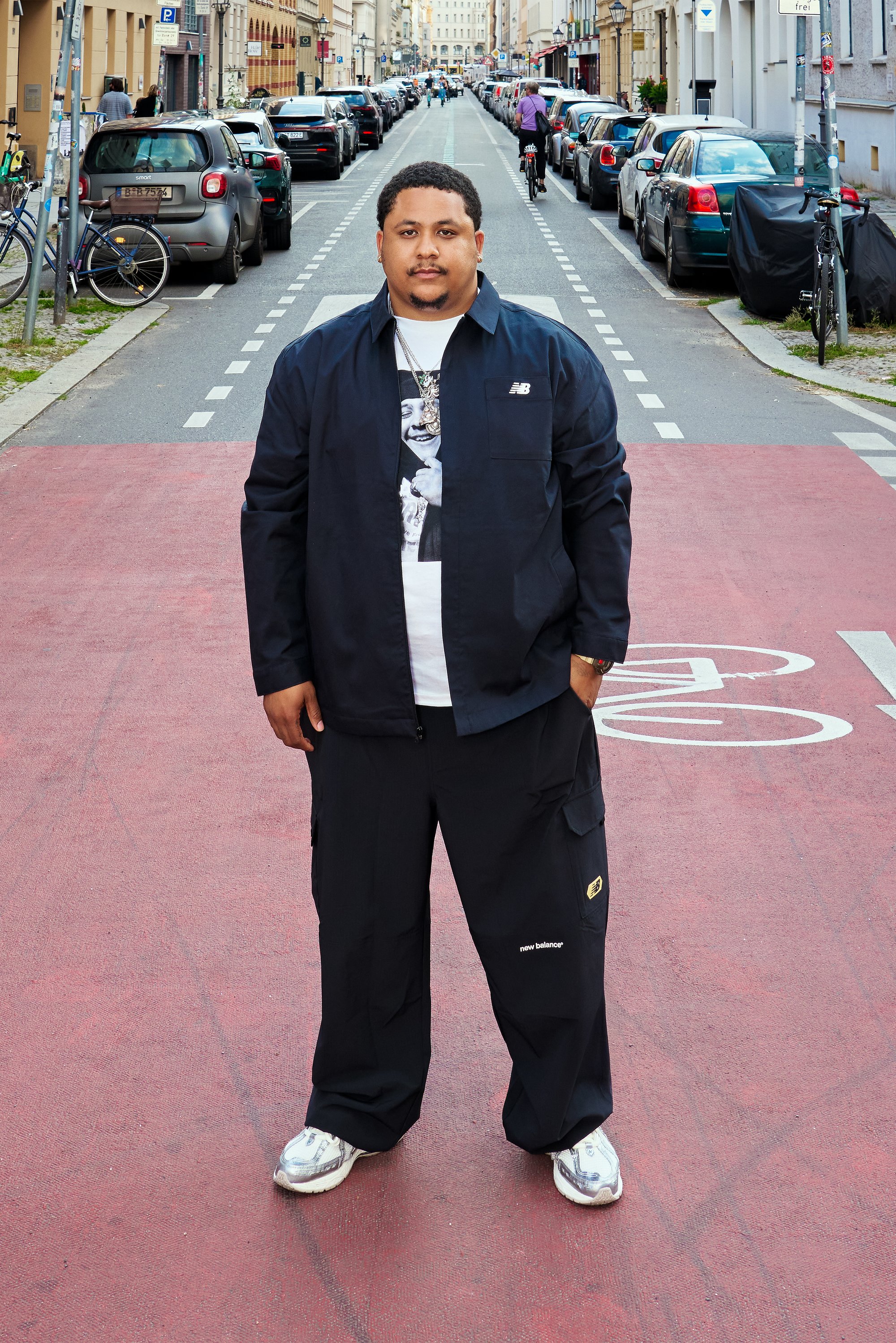 Mand i sort New Balance jakke og bukser, der står på en rød cykelbane i en bygade med parkerede biler og cyklister.