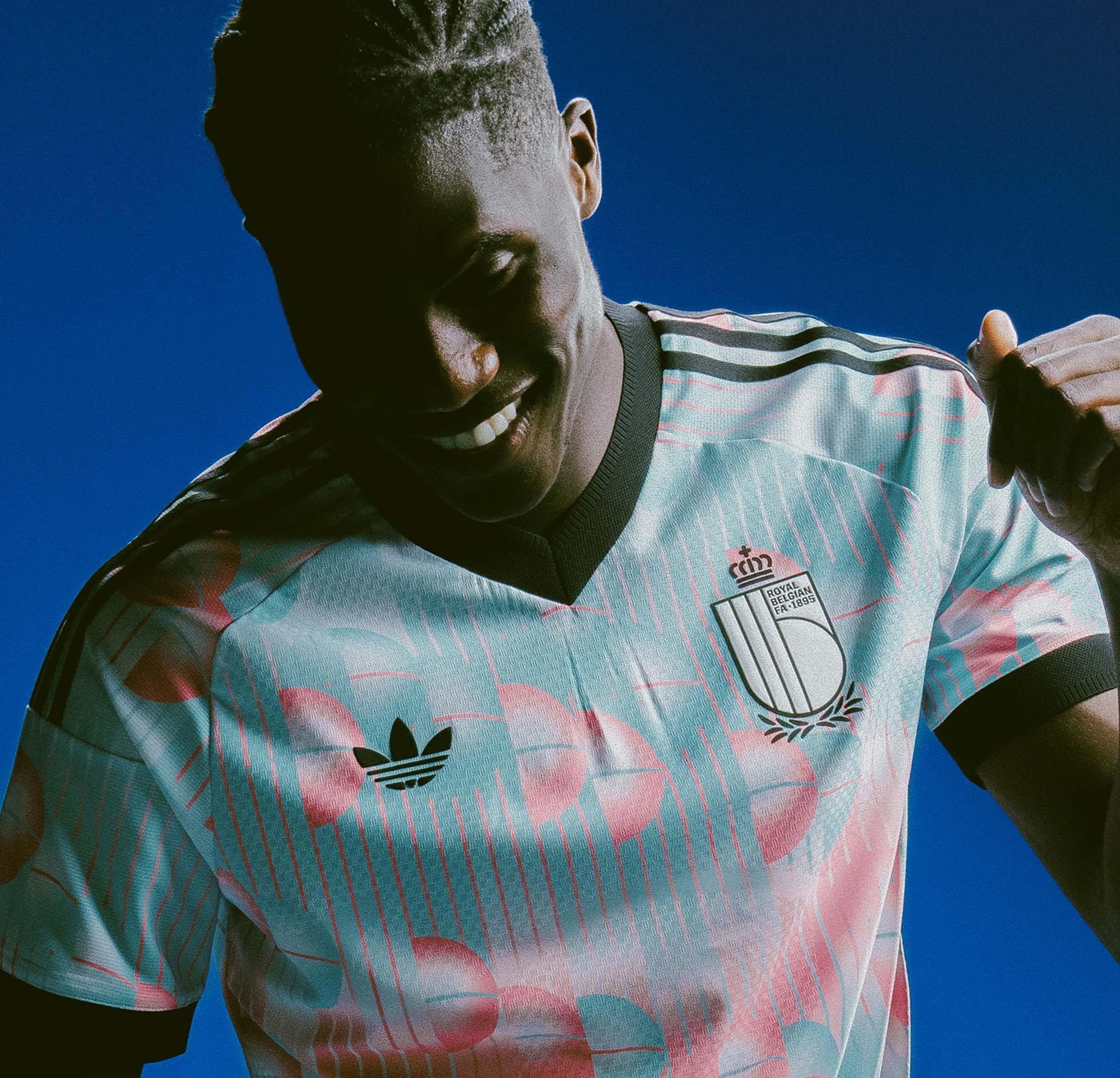 Man draagt een blauw en roze patroon Royal Belgian voetbaltrui tegen een heldere blauwe lucht, lachend en zijn arm opheffend.