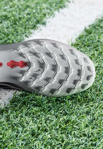 Semelle d'une chaussure de football blanche avec des crampons rouges posée sur un gazon artificiel vert près d'une ligne blanche peinte sur le terrain.