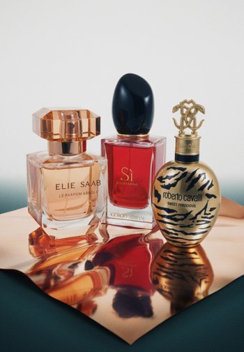 Three perfume bottles on a reflective copper surface: Elie Saab peach, Giorgio Armani red with black cap, and gold Roberto Cavalli with black animal print.