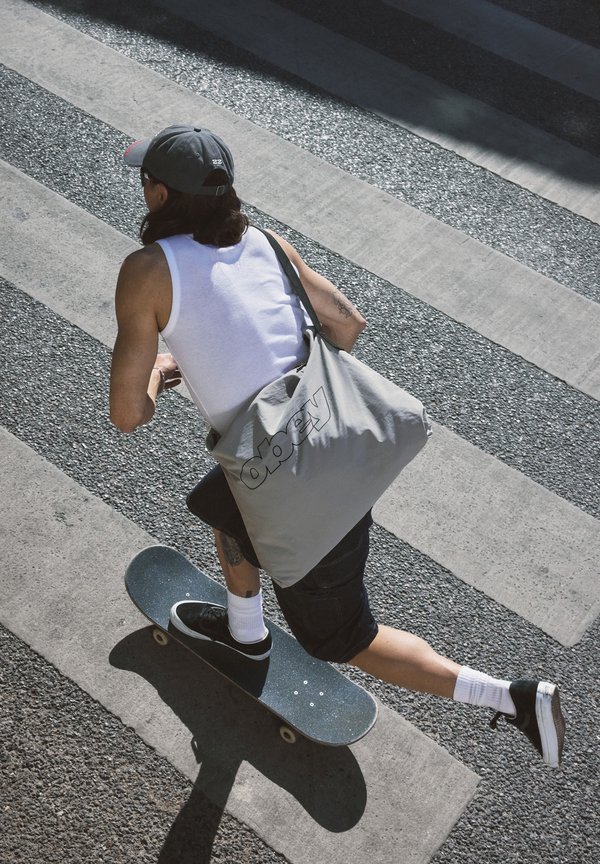 Skateboarder wearing a white tank top and cap, carrying a grey Obey bag, pushing off on a textured crosswalk in daylight.