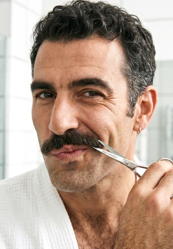 Homme aux cheveux bouclés et foncés taille une moustache épaisse avec de petits ciseaux dans une salle de bain lumineuse.