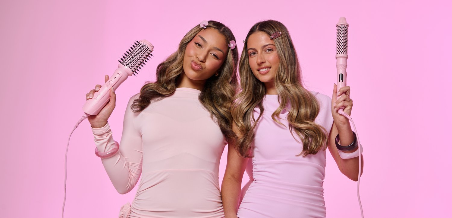 Deux femmes aux longs cheveux ondulés, vêtues de tenues rose clair, tiennent des brosses chauffantes roses devant un fond rose uni.