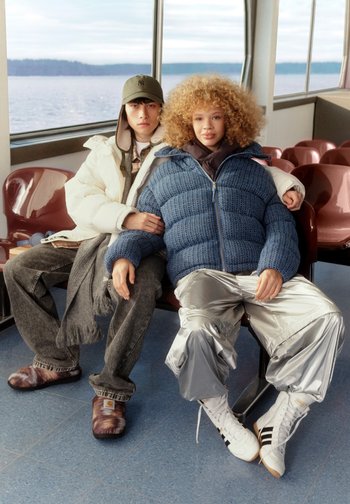 Two people sit on a bench. One wears a white padded jacket, grey scarf, dark jeans, and brown patterned slippers. The other has a blue padded jacket, silver trousers, and white high-top trainers.