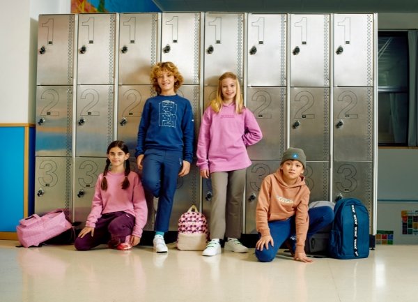 Gruppe på fire barn i fritidsklær: rosa og blå hettegensere, brune og grå bukser. Sølvfargede skap i bakgrunnen. Fargerike ryggsekker til stede.
