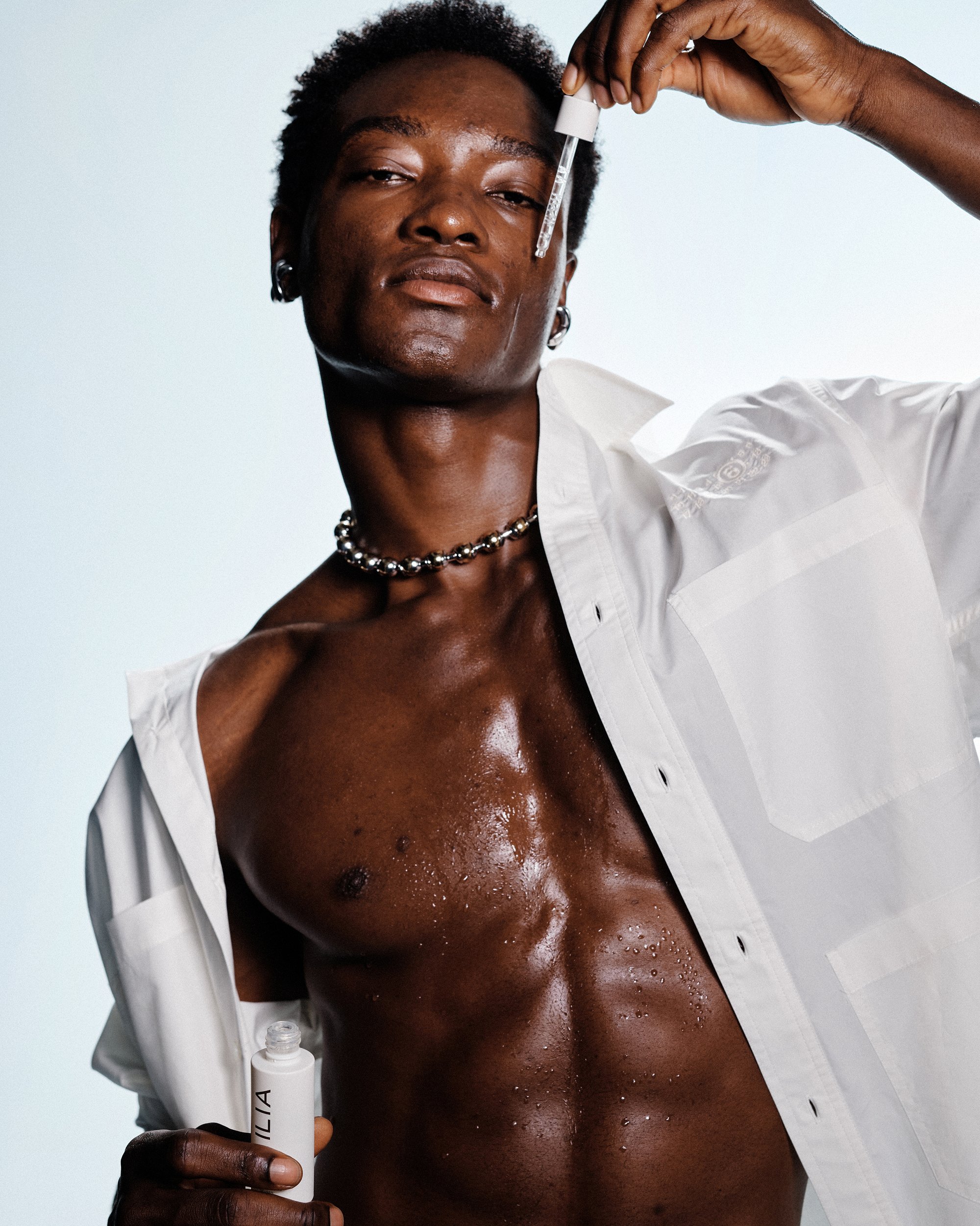 Een man met een gespierde, glanzende torso houdt een pipet boven zijn gezicht, terwijl hij een gedeeltelijk open wit shirt en een kralenketting draagt.