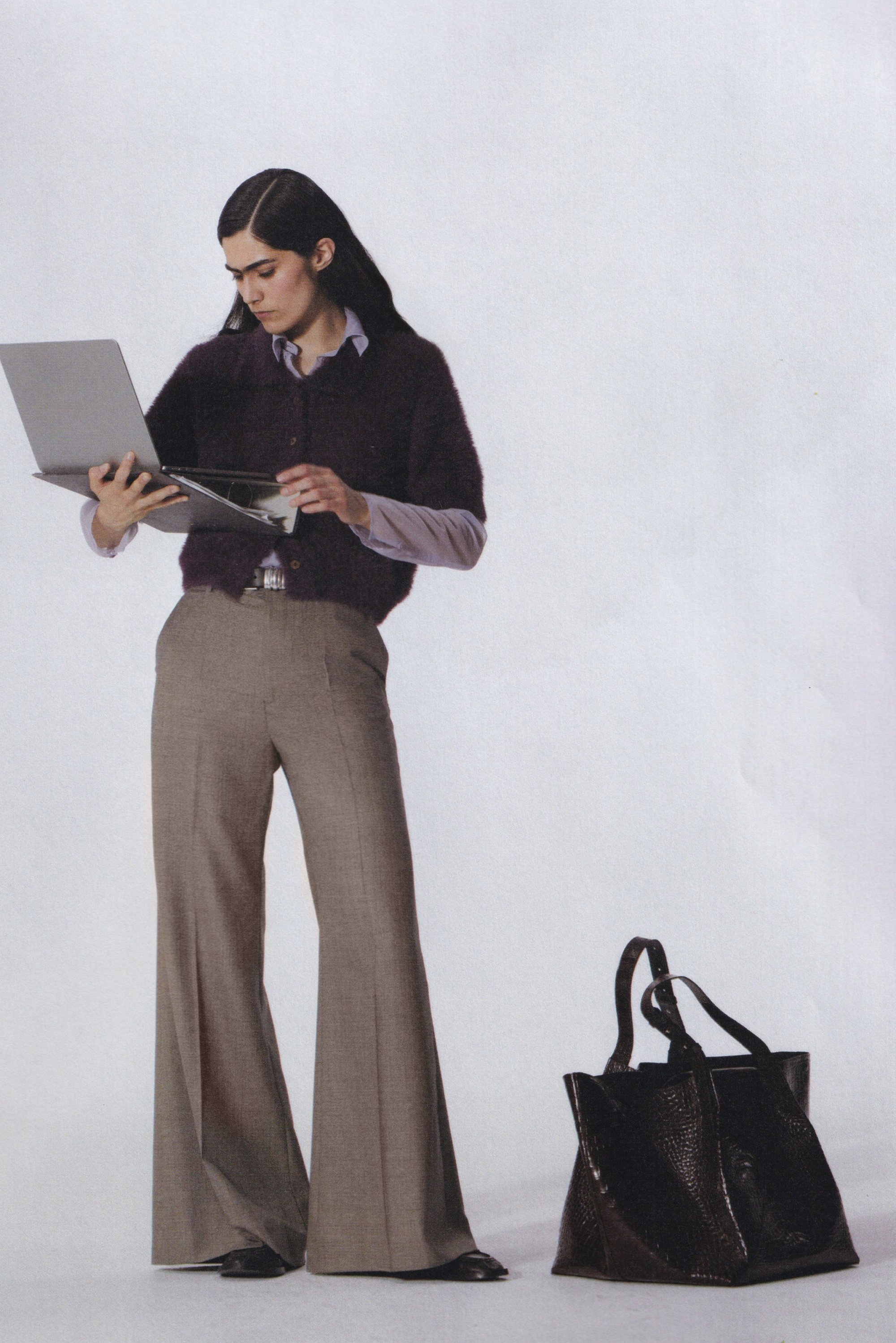 Grijze wollen broeken, donkerviolet fuzzy trui, lichtblauw shirt eronder, met een laptop in de hand. Zwarte gestructureerde handtas op de vloer.