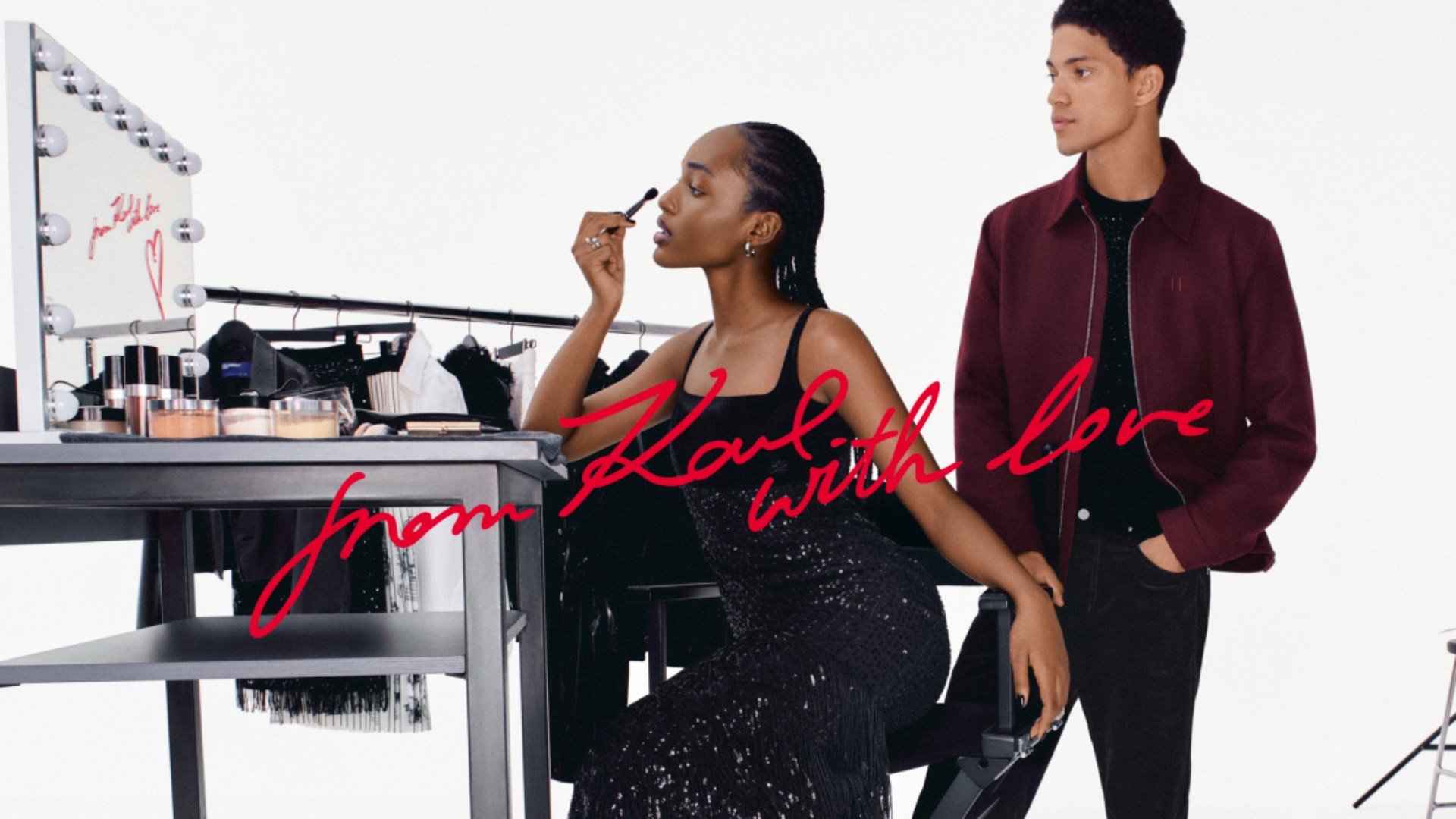 Schwarzes Paillettenkleid an einer Frau, die sich an einem Schminktisch schminkt, daneben steht ein Mann in bordeauxrotem Jackett, Spiegel und Kosmetikartikel sichtbar.