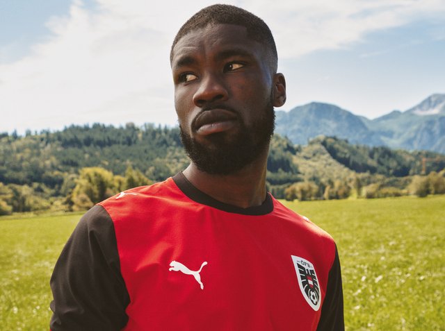 Ein Mann in einem roten und schwarzen Puma-Sporttrikot steht im Freien mit Bäumen und Bergen im Hintergrund und schaut nach links.