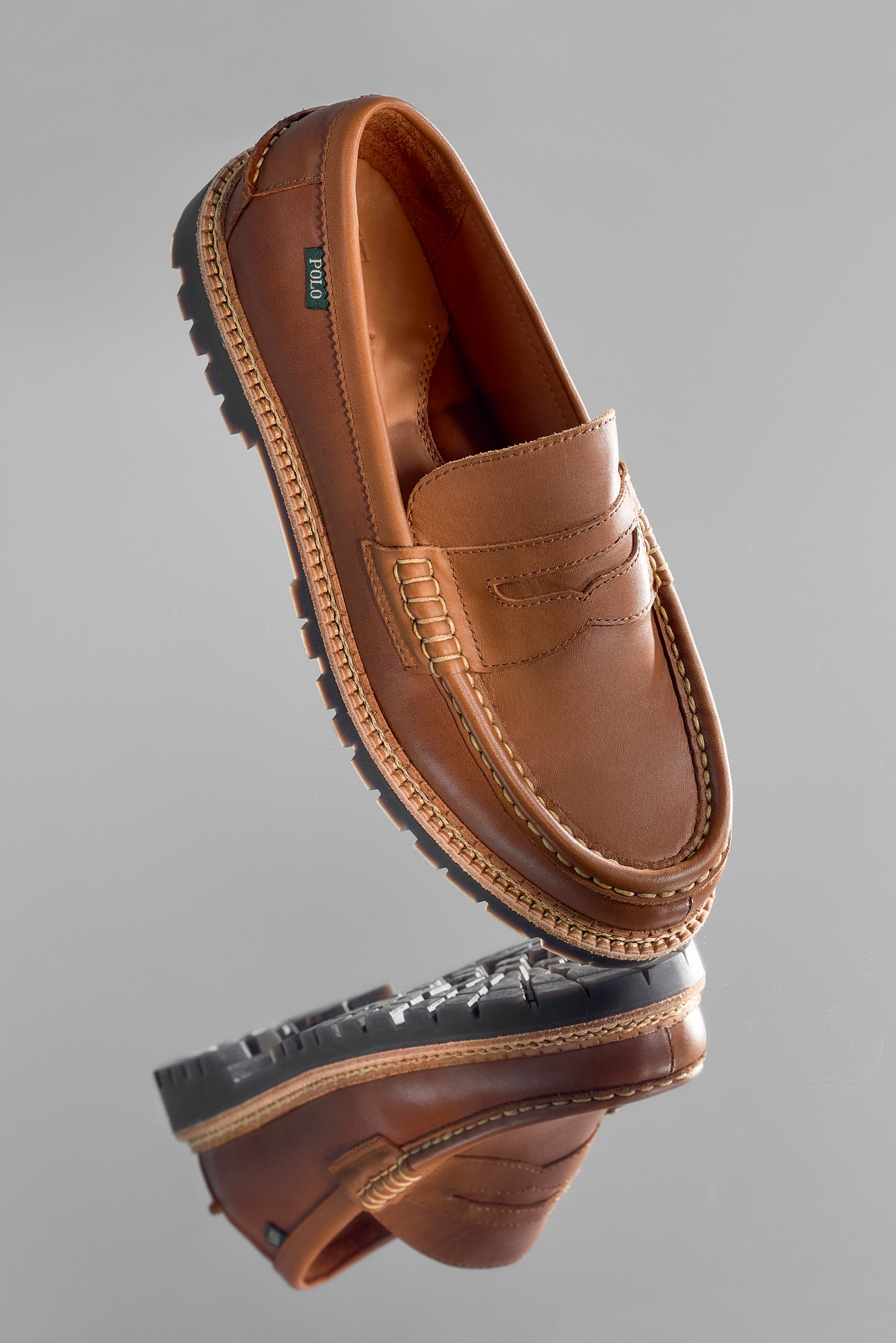Brown leather loafer shoe positioned upright, reflecting on a smooth surface. Detailed stitching and a textured sole are visible.