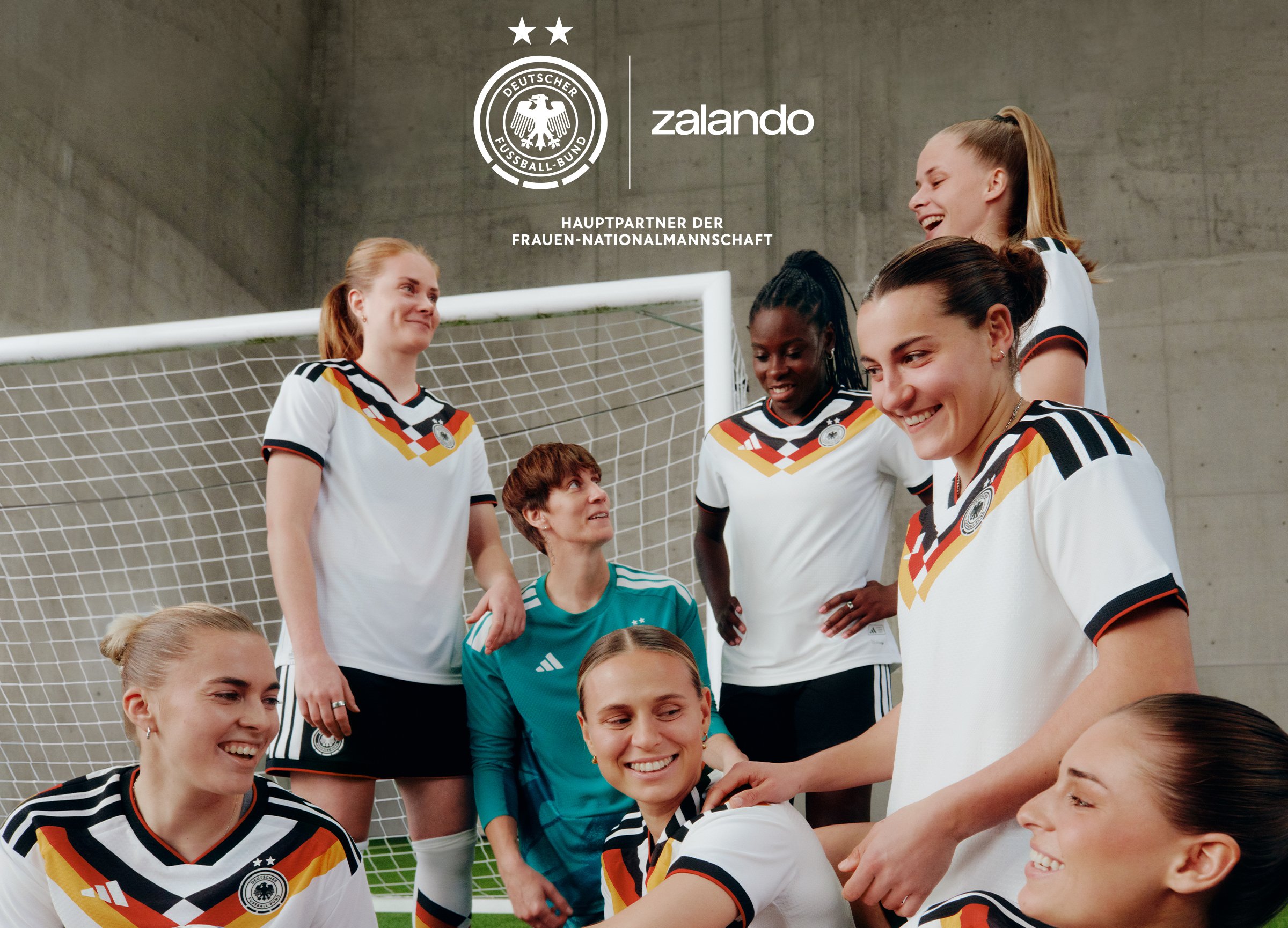 Sieben Fußballspielerinnen in Trikots der deutschen Nationalmannschaft lächeln und interagieren in der Nähe eines Torpfostens in der Halle.