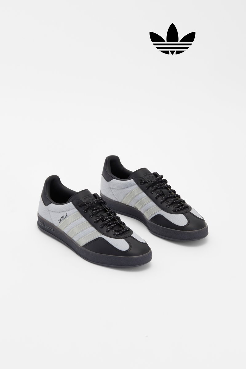 Zapatillas deportivas negras y grises con cordones, que presentan tres franjas blancas y el logo "Gazelle", exhibidas sobre un fondo blanco liso.