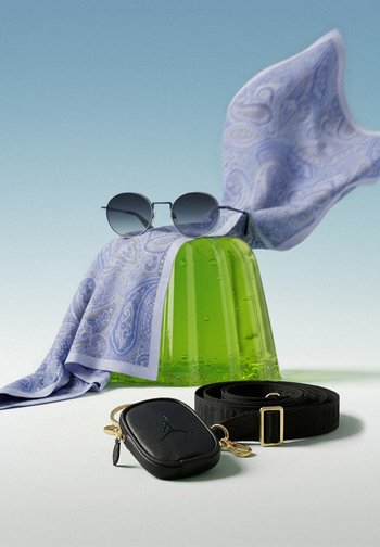 Green translucent jelly shoes with a blue paisley scarf and round sunglasses placed on top, a black leather pouch with a strap in front on a white surface.