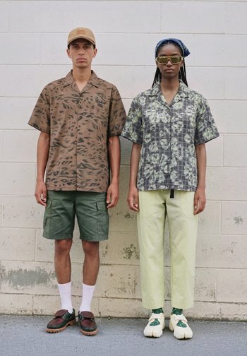 Two people stand side by side against a wall, wearing patterned shirts, casual pants, and accessories like a cap, sunglasses, and headscarf.