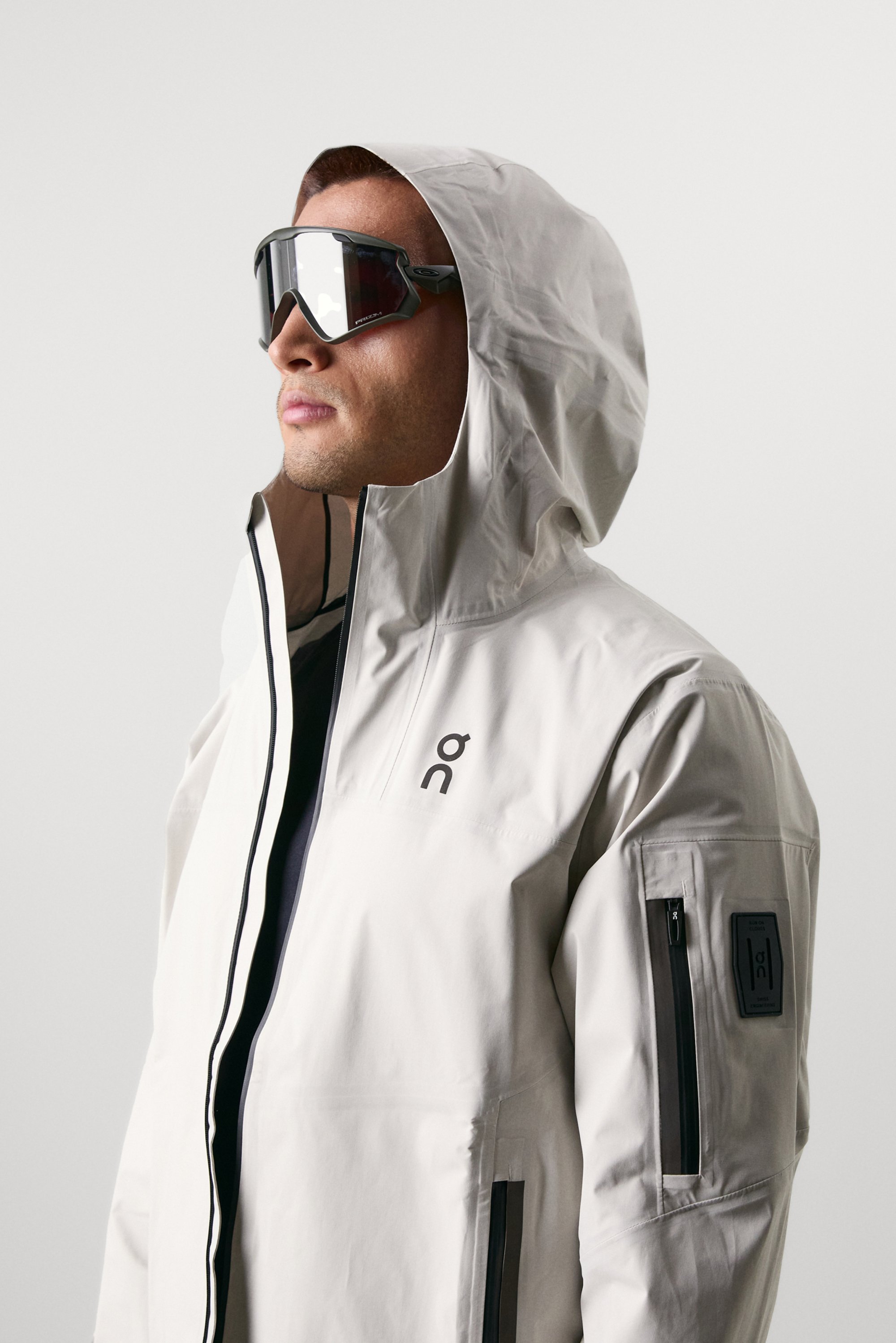 A man wearing a light-colored waterproof jacket with a hood and large sunglasses, facing slightly to his left against a plain background.
