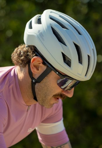 Homme portant un casque de vélo blanc, des lunettes de cyclisme miroir et un maillot rose avec un brassard blanc, penché en avant en extérieur.