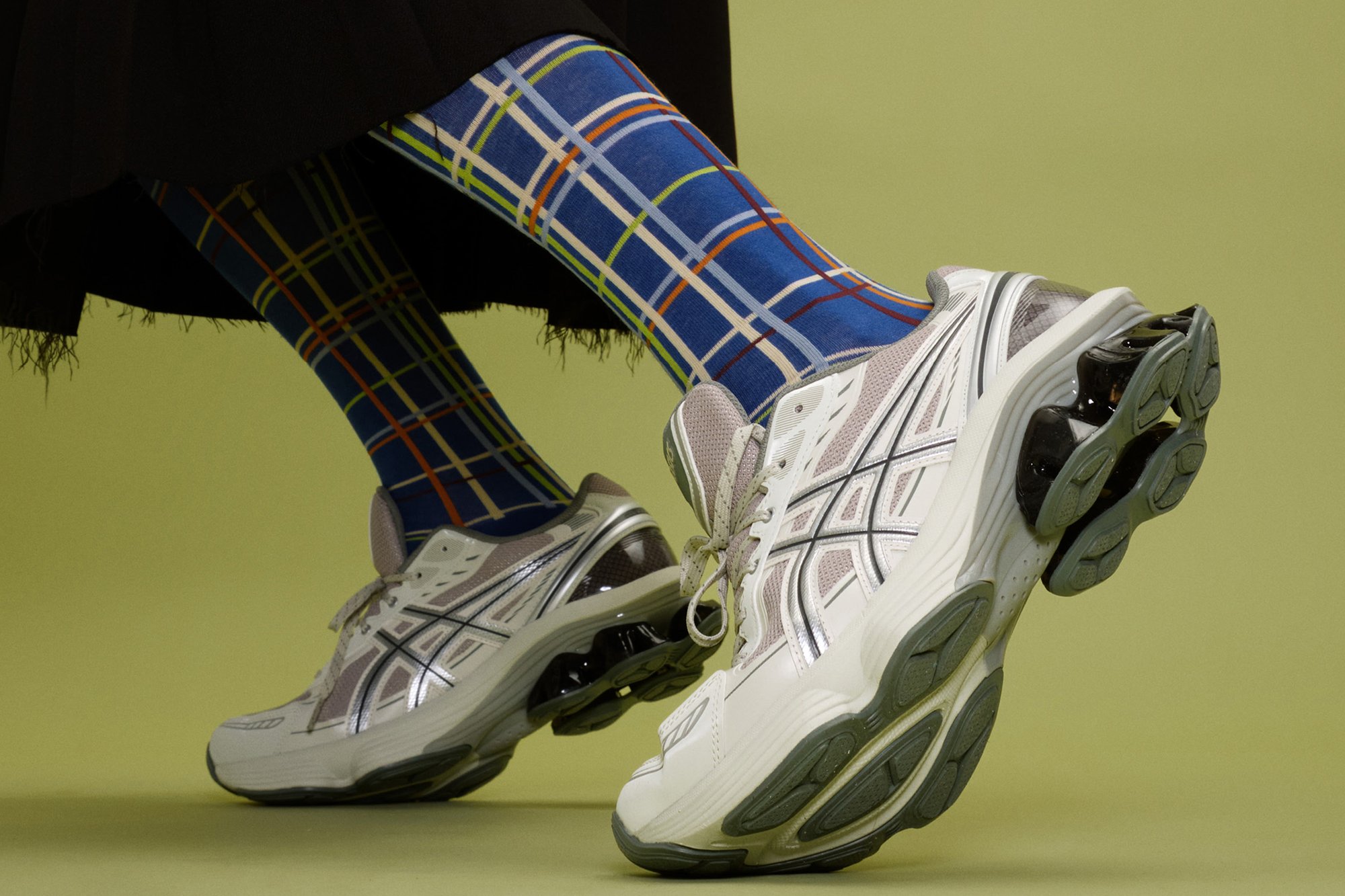 Zapatillas de running Asics en blanco con detalles plateados y suelas de un gris texturizado, combinadas con calcetines azules que presentan un colorido patrón de cuadros.