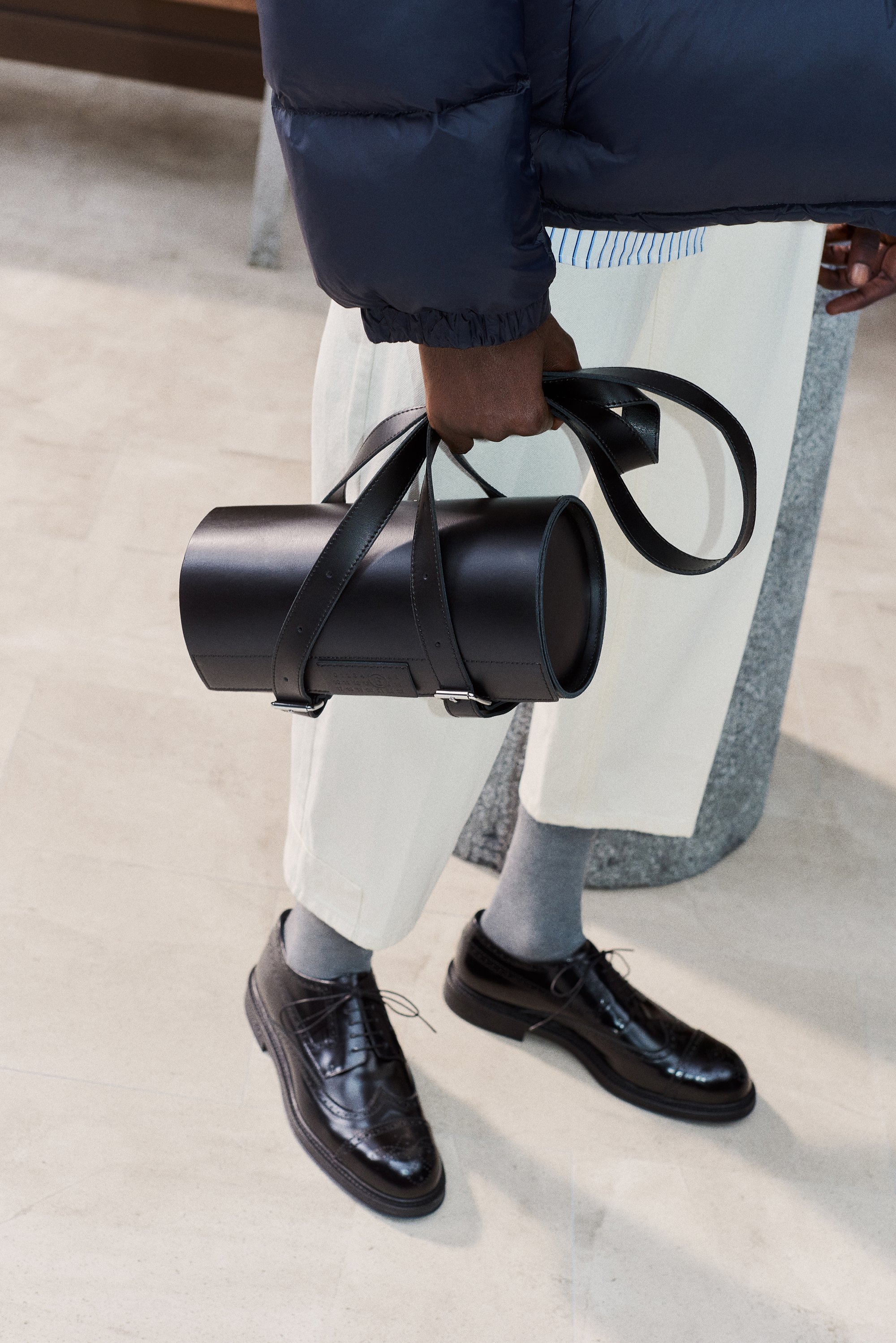 Person in a navy puffer jacket holds a cylindrical black bag with leather straps, wearing light pants and polished black shoes.