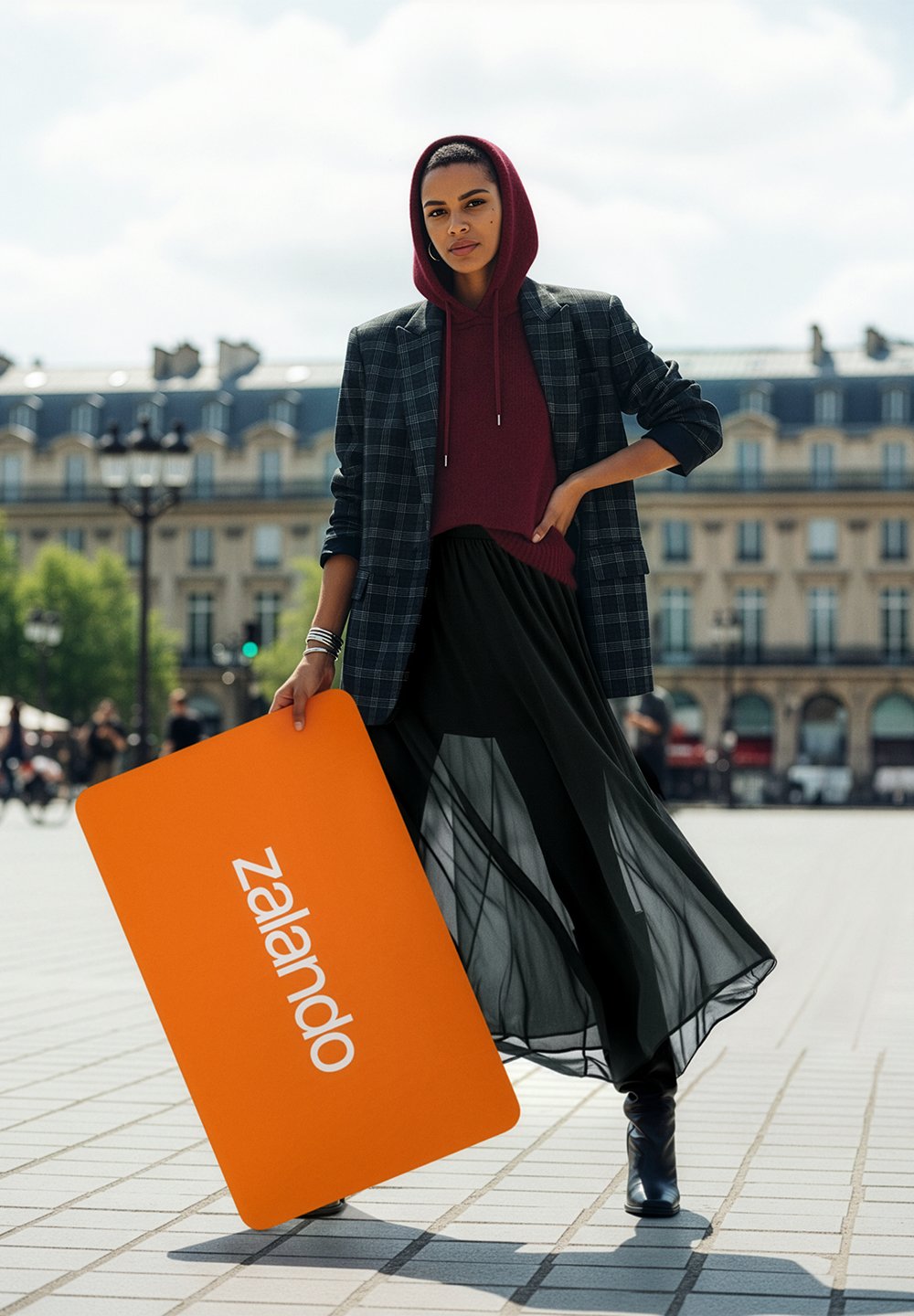 Modell in einem karierten Blazer über einem burgunderroten Kapuzenpullover, kombiniert mit einem schwarzen, transparenten Rock. Hält ein orangefarbenes, rechteckiges Zalando-Schild.