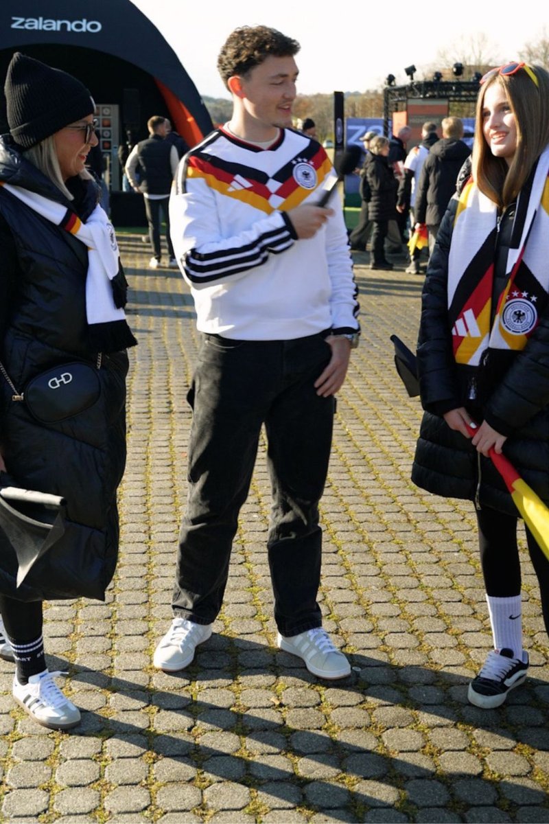 Three people wearing German soccer scarves and jackets stand outside near a Zalando tent, engaging in conversation on a sunny day.