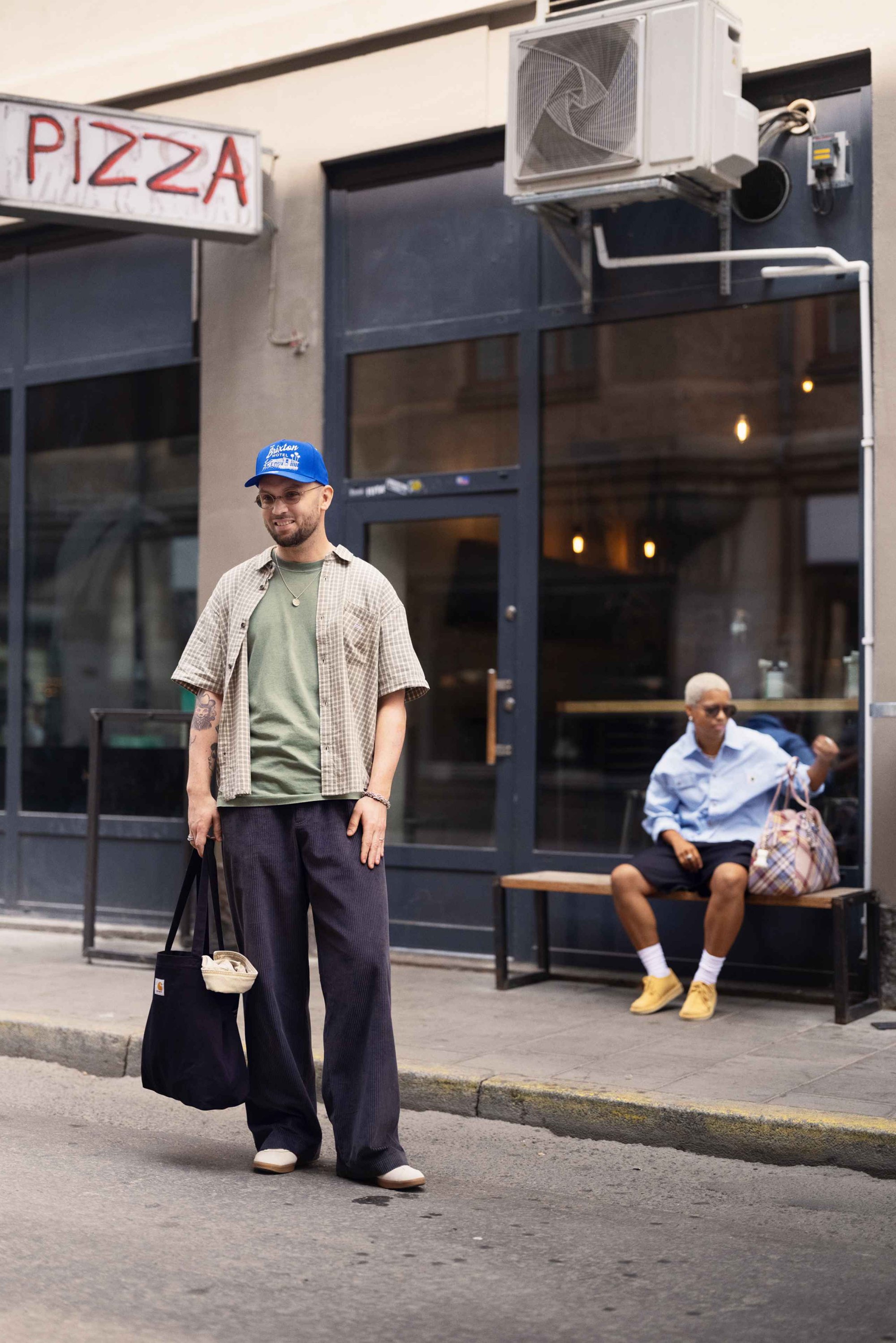 Informele outfit met een oversized geruit kortemouwen shirt, groene T-shirt, donkere wijde broek, blauwe pet en zwarte draagtas.