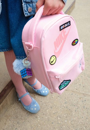 Enfant tenant un sac à dos Nike rose avec des écussons colorés et un porte-clés, portant des chaussures bleues, des collants roses et une jupe en jean.
