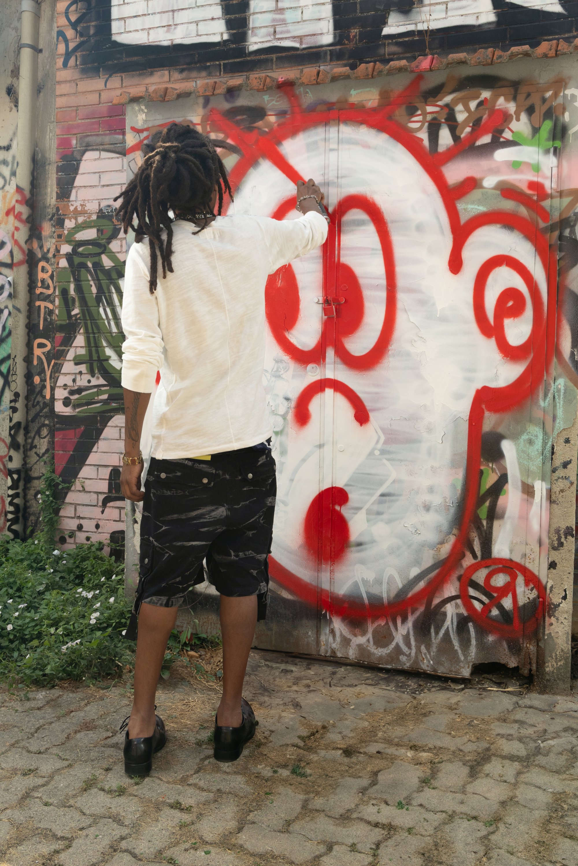 Persona con dreadlocks largos, vistiendo una camiseta blanca de manga larga y pantalones cortos negros con estampados, pinta un diseño abstracto rojo en una puerta cubierta de graffiti.