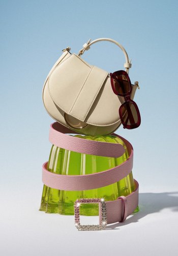 Cream half-moon handbag with dark reddish-brown sunglasses, a light pink belt with a rhinestone buckle wrapped around a green translucent mould.