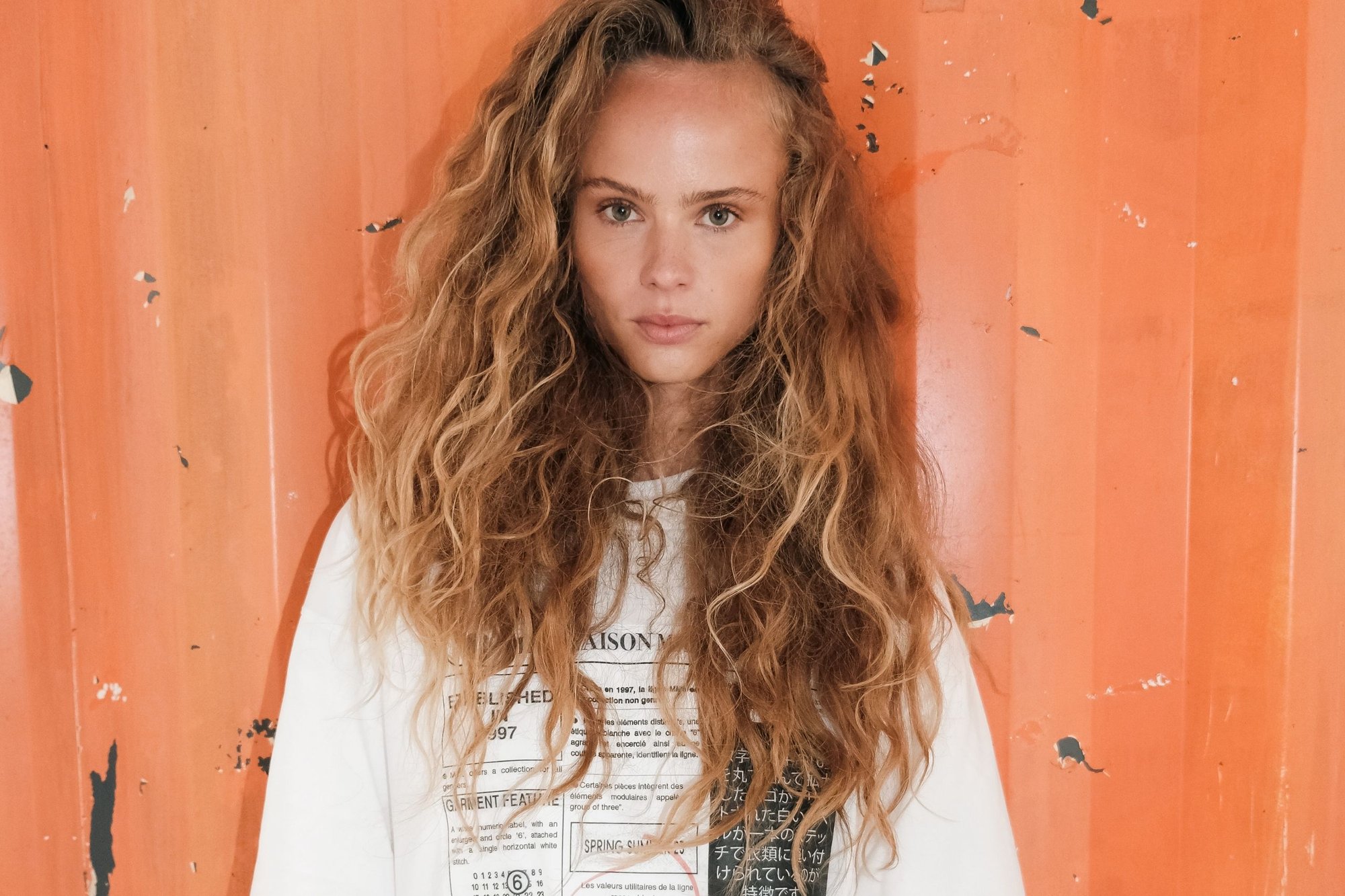 Jonge vrouw met lang krullend haar staat tegen een versleten oranje metalen muur, draagt een wit graphic sweatshirt en kijkt recht vooruit.